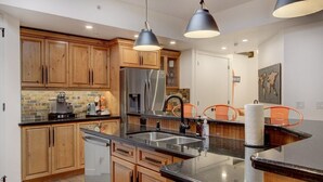 A modern kitchen with wooden cabinets, a stainless steel refrigerator, a coffee machine, and a large island with a sink. - Mistaya Suite | Stylish Downtown Penthouse (Canmore)