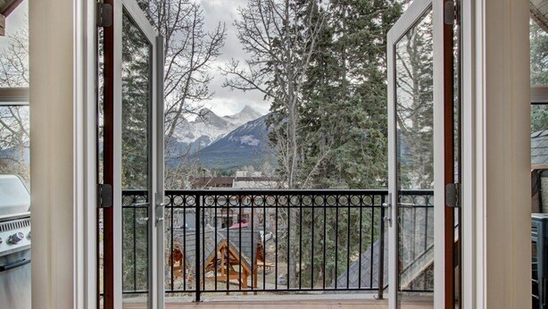 A deck with a metal railing, wooden flooring, and a view of mountains and trees. - Mistaya Suite | Stylish Downtown Penthouse (Canmore)