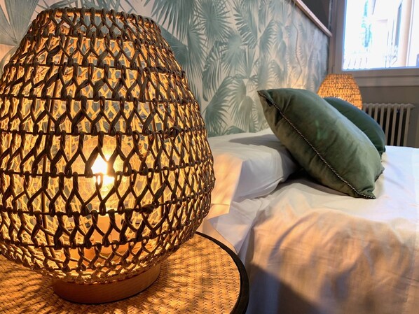 A room with a woven lamp on a woven base, a green pillow on the bed, and a window with a view of palm trees. - Exclusive accommodations in downtown Burgos. (Burgos)