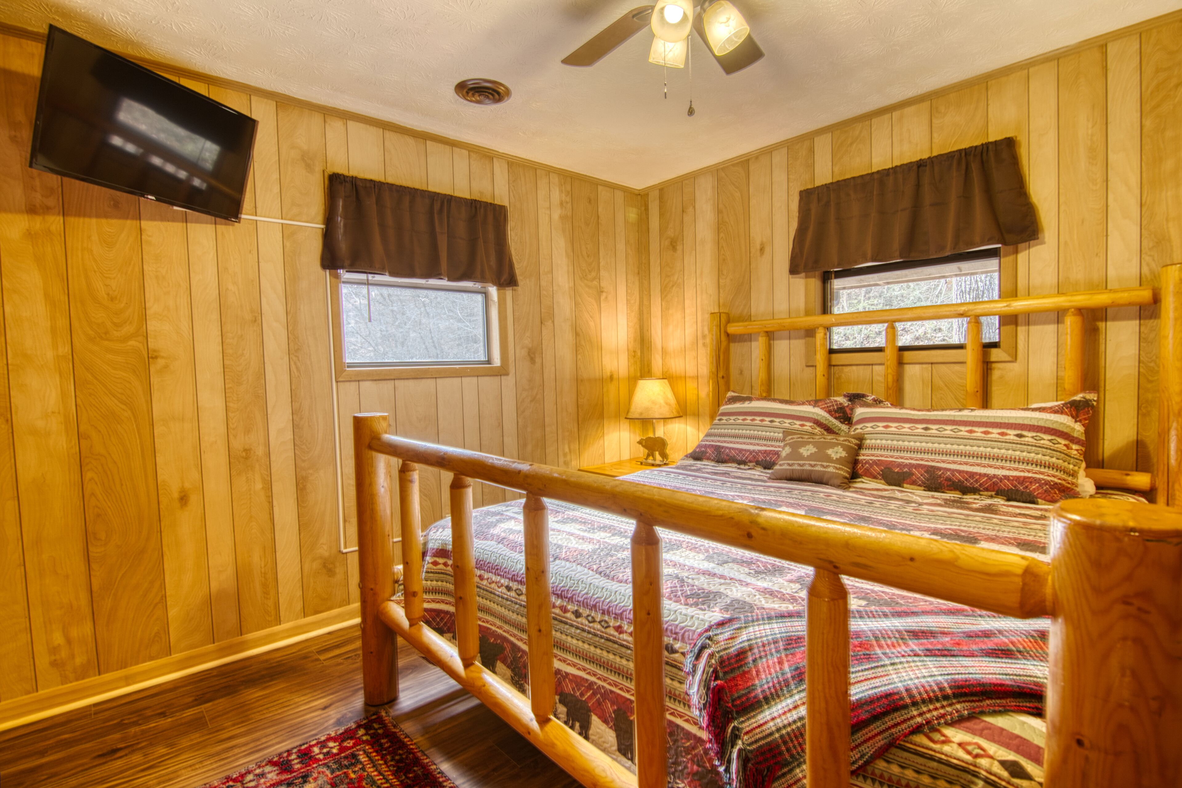 A wooden-paneled room with a bed, a TV mounted on the wall, and a ceiling fan.