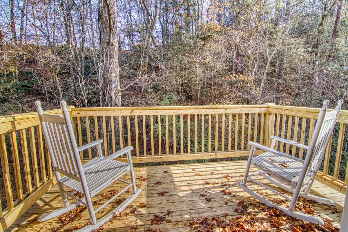 A wooden deck with two metal rocking chairs, surrounded by trees and fallen leaves.