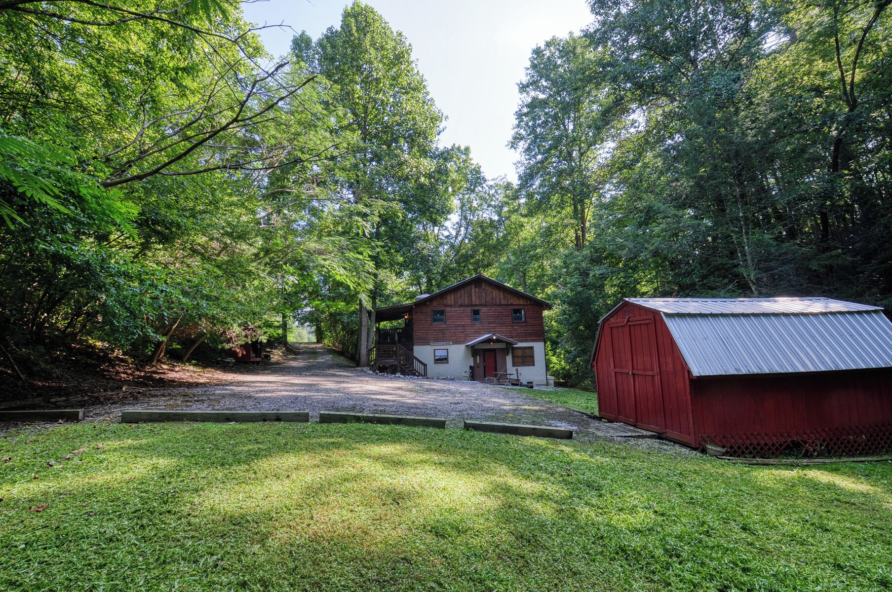 A rustic cabin with a wooden exterior and a metal roof, surrounded by lush greenery and a gravel path leading to the entrance.