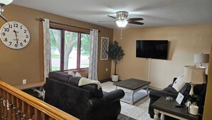 A living room with a large clock, a ceiling fan, a flat-screen TV, and a staircase.