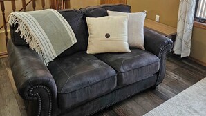 A living room with a black leather sofa, a wooden door, a wall clock, and a window with curtains.