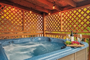 Ein Whirlpool mit Holzterrasse und Gitterwänden, eine Weinflasche und ein Tablett mit Trauben und Gläsern.
