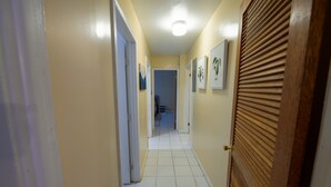 Un pasillo estrecho con suelo de baldosas blancas y paredes color beige, que conduce a una habitación con una puerta de madera y una lámpara en el techo.