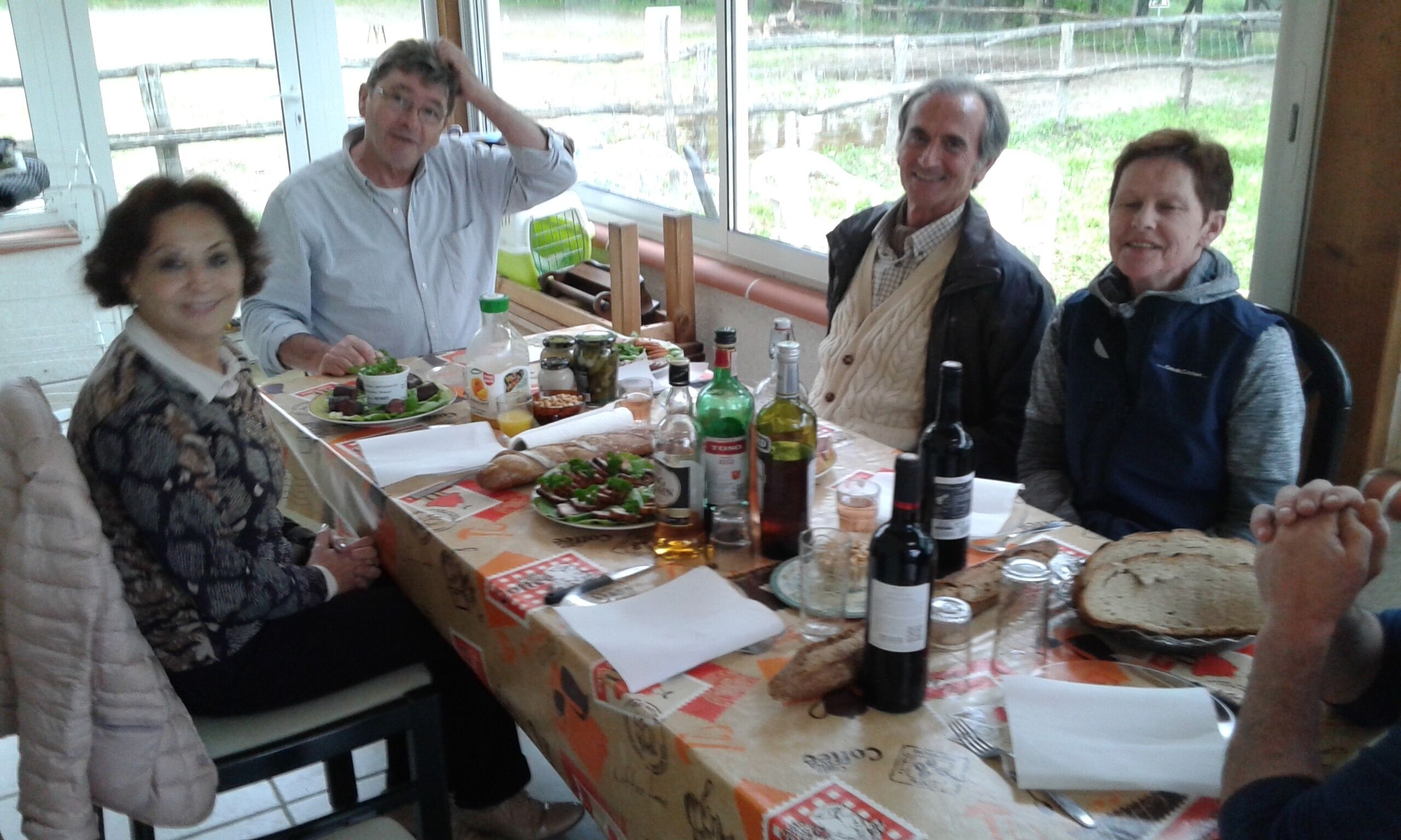 Een groep mensen zit rond een tafel met eten en drinken, er is een raam zichtbaar en een uitzicht op het landschap.