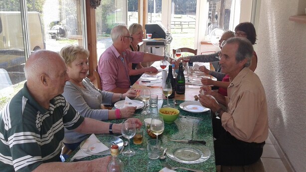 Een groep mensen zit aan tafel met verschillende gerechten en drankjes, wat wijst op een gezellig samenzijn of maaltijd.