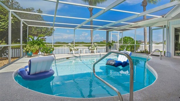 A covered outdoor swimming pool with clear blue water, surrounded by a white fence and palm trees. - Oasis on the Gulf, screened pool, sunset, kayaks! (Hernando Beach)