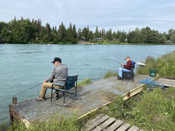 Two individuals fishing by a calm river with a dock and chairs. - LOCATED ON THE KENAI RIVER!!  Kenai River Haven Unit #4. (Soldotna)