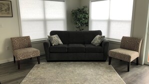 Ein Wohnzimmer mit einem dunkelgrauen Sofa, zwei gemusterten Sesseln, einem großen beigen Teppich und einer Topfpflanze.
