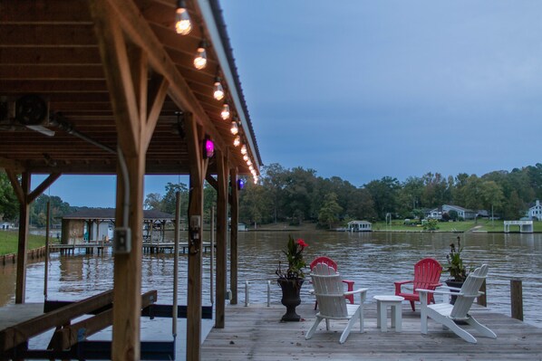 Un muelle de madera con sillas y una mesa, con vistas a un lago tranquilo y casas al fondo.