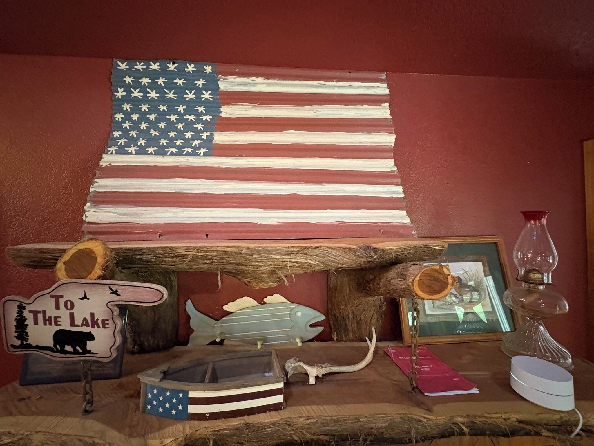 A rustic interior with a large American flag, a wooden sign reading "To The Lake", a framed picture, a glass vase, and a small boat model.