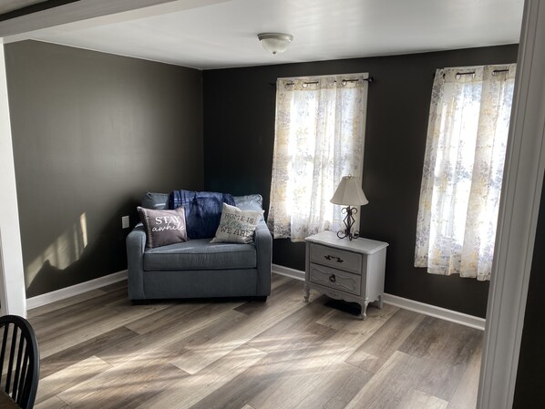 A room with a grey sofa, a white nightstand, and a lamp. There are two windows with floral curtains. - Cozy Country farmhouse with Hot Tub (Meshoppen)