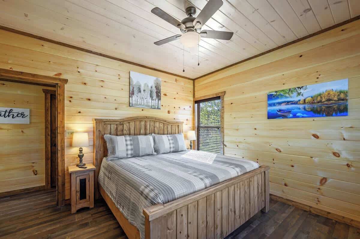 A wooden cabin bedroom with a bed, a nightstand, a ceiling fan, and two paintings on the walls.