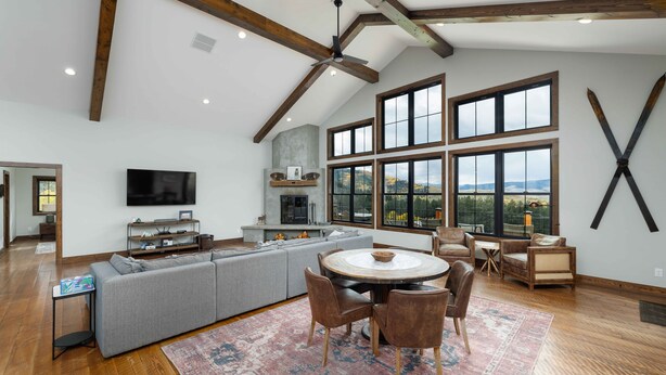 A spacious living area with a large gray sectional sofa, a round dining table with chairs, and a TV mounted on the wall.