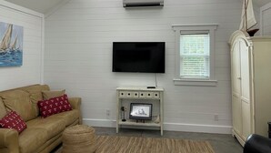 A living room with a flat-screen TV mounted on the wall, a ceiling fan, a window with blinds, a white cabinet, and a beige sofa with patterned cushions. - Walking distance to Downtown Beaufort, NC. Short drive to several beaches. (Beaufort)