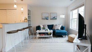 Un salon moderne avec un canapé bleu, une table basse blanche et un téléviseur posé sur un meuble. On y trouve également un coin cuisine avec un bar et des tabourets.