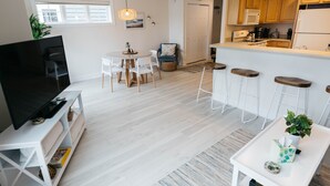 Un salon moderne doté d’un téléviseur à écran plat, d’un coin repas avec une table ronde et des chaises, d’une cuisine avec des meubles en bois et des tabourets de bar, et d’un petit espace détente agrémenté d’une plante.