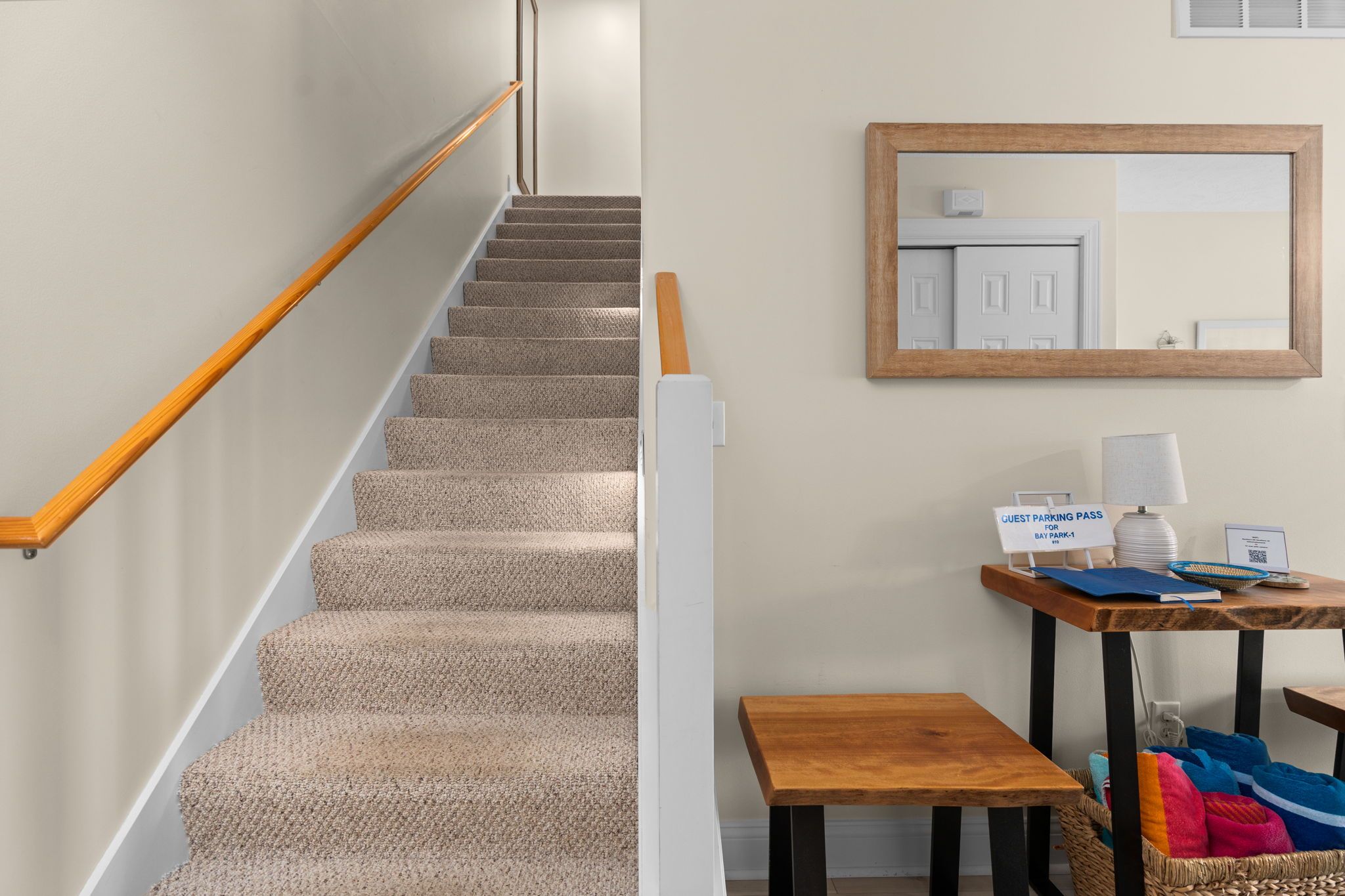 A carpeted staircase with a wooden handrail, a wall-mounted mirror, a table with a guest parking pass, and a basket with colorful items.