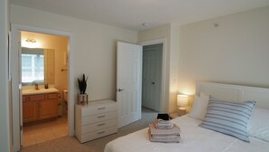 Une chambre à coucher comprenant un lit, une table de chevet avec une lampe, une commode, une coiffeuse, un miroir et une porte donnant sur une salle de bain.
