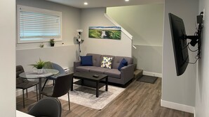 A modern living room with a grey sofa, a coffee table, and a dining area with a glass table and chairs. There is a painting on the wall and a staircase leading to another level. - The Hamm Suite (Saskatoon)
