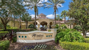 A landscaped entrance with a sign for Windsor Hills Clubhouse, surrounded by palm trees and well-maintained hedges. - Under the Sea Villa – 3mi to Disney! Remodeled! 5 Bed&Bath, Private Pool/Spa! (Four Corners)