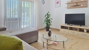 Uma sala de estar com sofá, mesa de centro de vidro, televisão sobre um suporte de madeira, planta em vaso e relógio na parede.