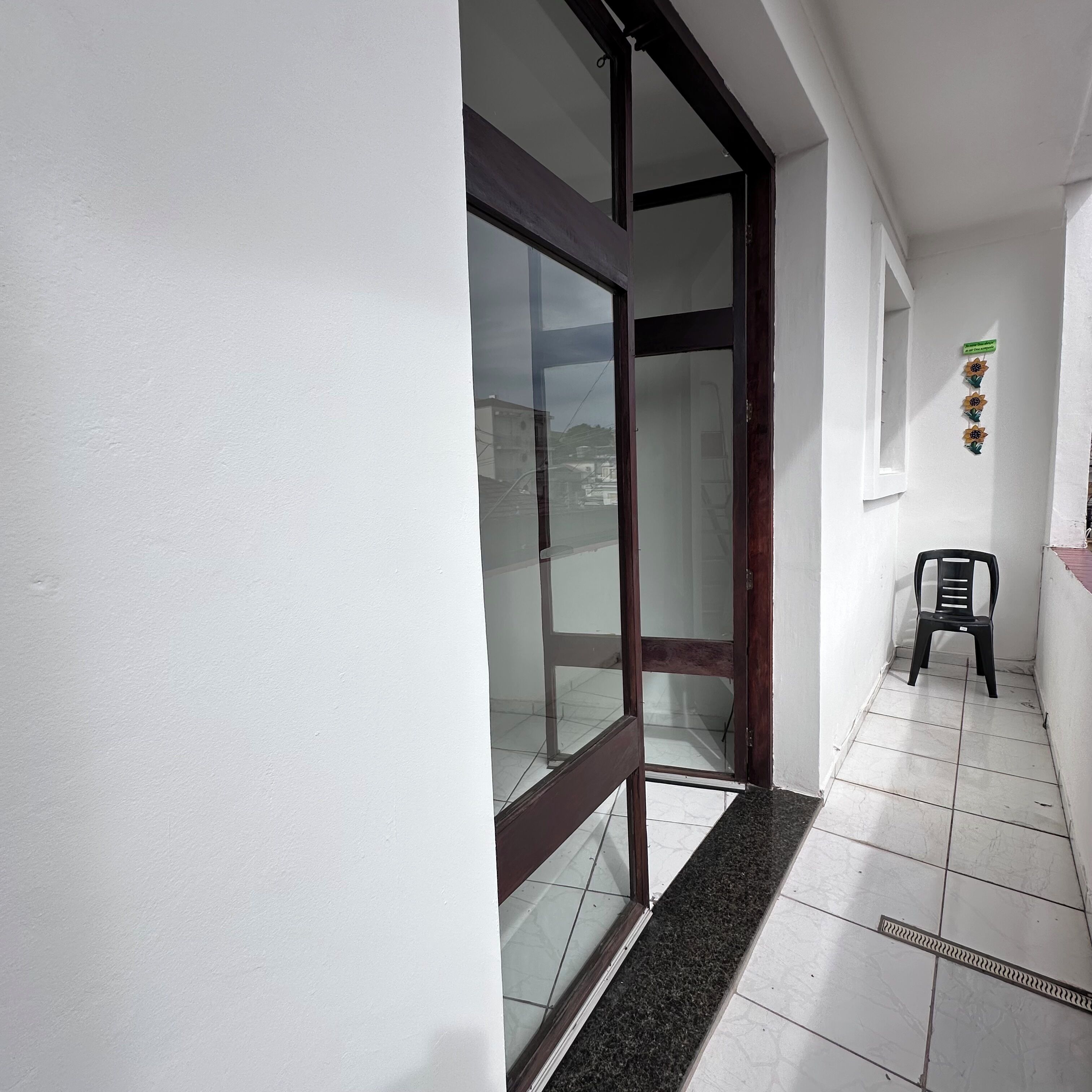 Um corredor com uma porta de vidro, uma cadeira de plástico preta e um chão ladrilhado.