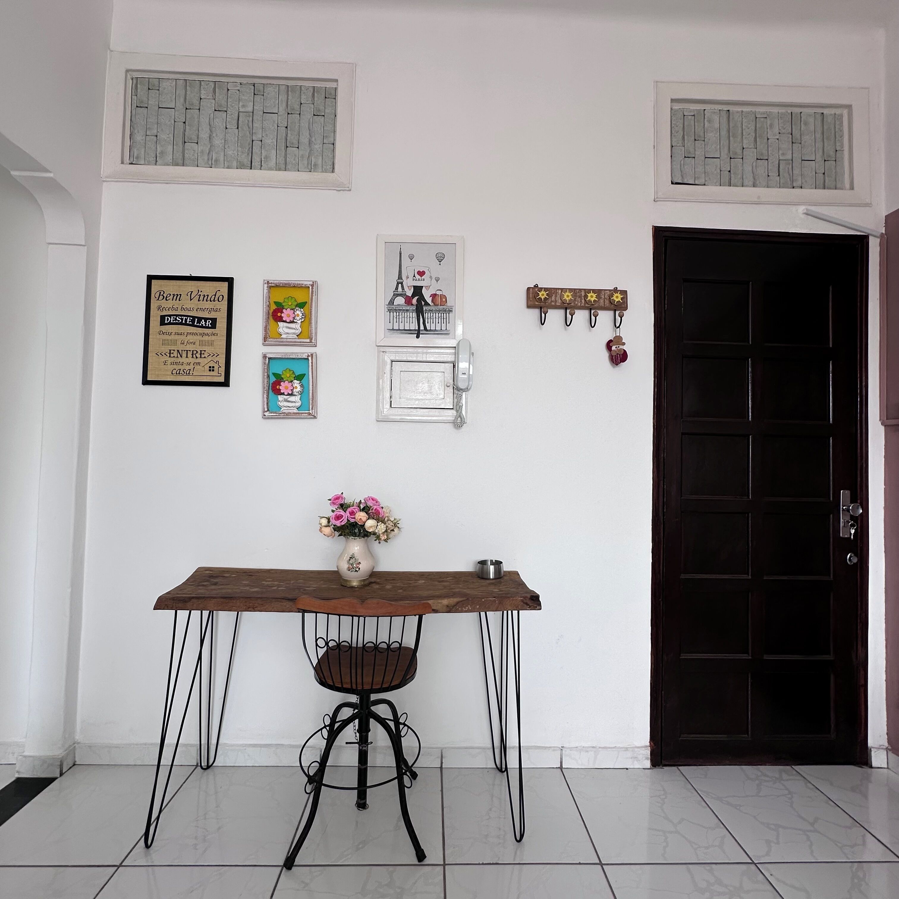 Um quarto com uma mesa de madeira, uma cadeira, um vaso com flores, um telefone na parede e uma porta.