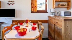 A small, cozy kitchen with wooden cabinets, a dining table set with red bowls and a basket of bread, and a window with curtains. - Nécou 505 by Interhome (Les Menuires)