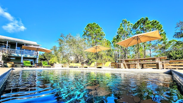 A clear swimming pool with lounge chairs and umbrellas, surrounded by trees and a building with a balcony. - 🌴Beachside Escape Private Modern Heated Pool - Vacation Getaway Gulf Shores🌴 (Gulf Shores)
