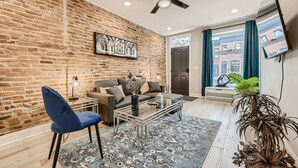 Un salon avec un mur en briques, un ventilateur de plafond, un canapé, une table basse et une grande fenêtre avec des rideaux.