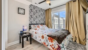 Une chambre à coucher avec un lit, une table de chevet, un ventilateur de plafond et une fenêtre avec des rideaux.