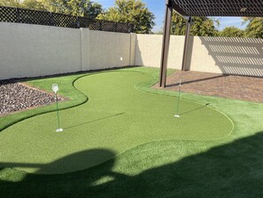 Un campo de golf con césped artificial bien cuidado, con dos hoyos, rodeado por un patio de ladrillo y una valla de madera.