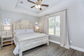 Une chambre avec un lit en bois, deux tables de chevet avec des lampes, un ventilateur de plafond, une grande fenêtre avec des rideaux et une vue sur l’extérieur.