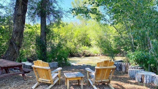 A rustic outdoor seating area with wooden chairs and a picnic table, surrounded by trees and a small stream.