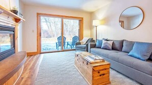 A modern living room with a grey sofa, a wooden coffee table, and a large window with a view of outdoor chairs.