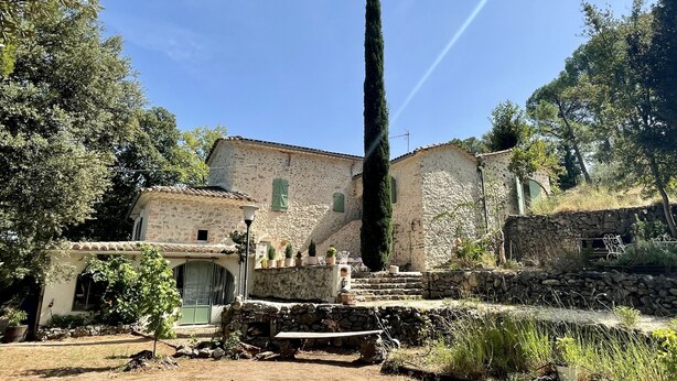A rustic stone building with a tall cypress tree in front, surrounded by greenery and a clear sky.