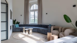 Ein modernes Wohnzimmer mit einem gebogenen grauen Sofa, einem runden Couchtisch und einem beigen Sessel. Zwei Holznachttische, eine große Zimmerpflanze und ein Blick ins Freie durch ein Fenster mit beigen Vorhängen.