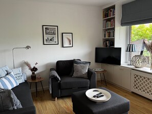 A modern living room with a sofa, a coffee table with remotes, a bookshelf, a TV, and a window with a view of greenery.