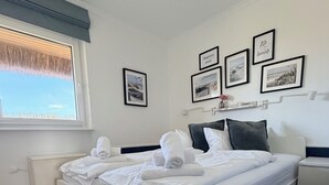 A neatly made bed with white linens and towels, a bedside table with a lamp, a window with a view of the sky, and framed pictures on the wall. - PiPPA - PIPPA, Germany (List)