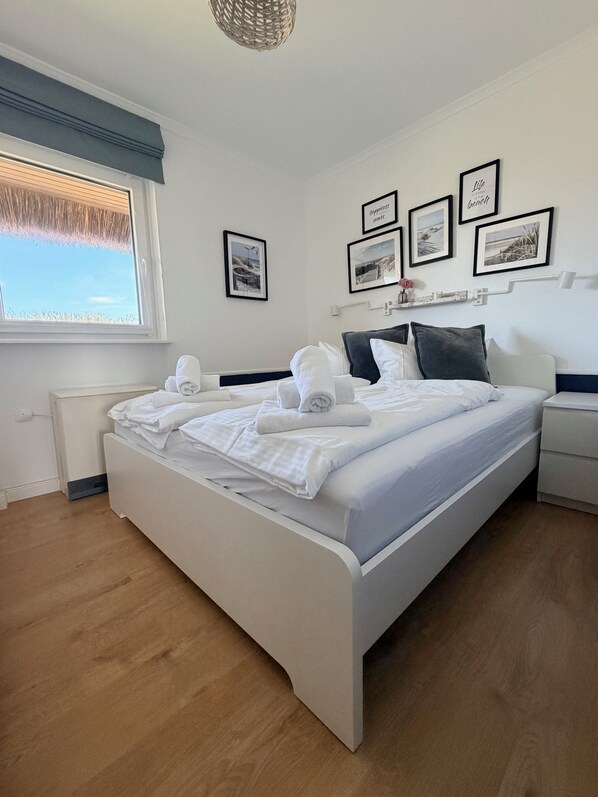 A neatly made bed with white linens and towels, a bedside table with a lamp, a window with a view of the sky, and framed pictures on the wall.