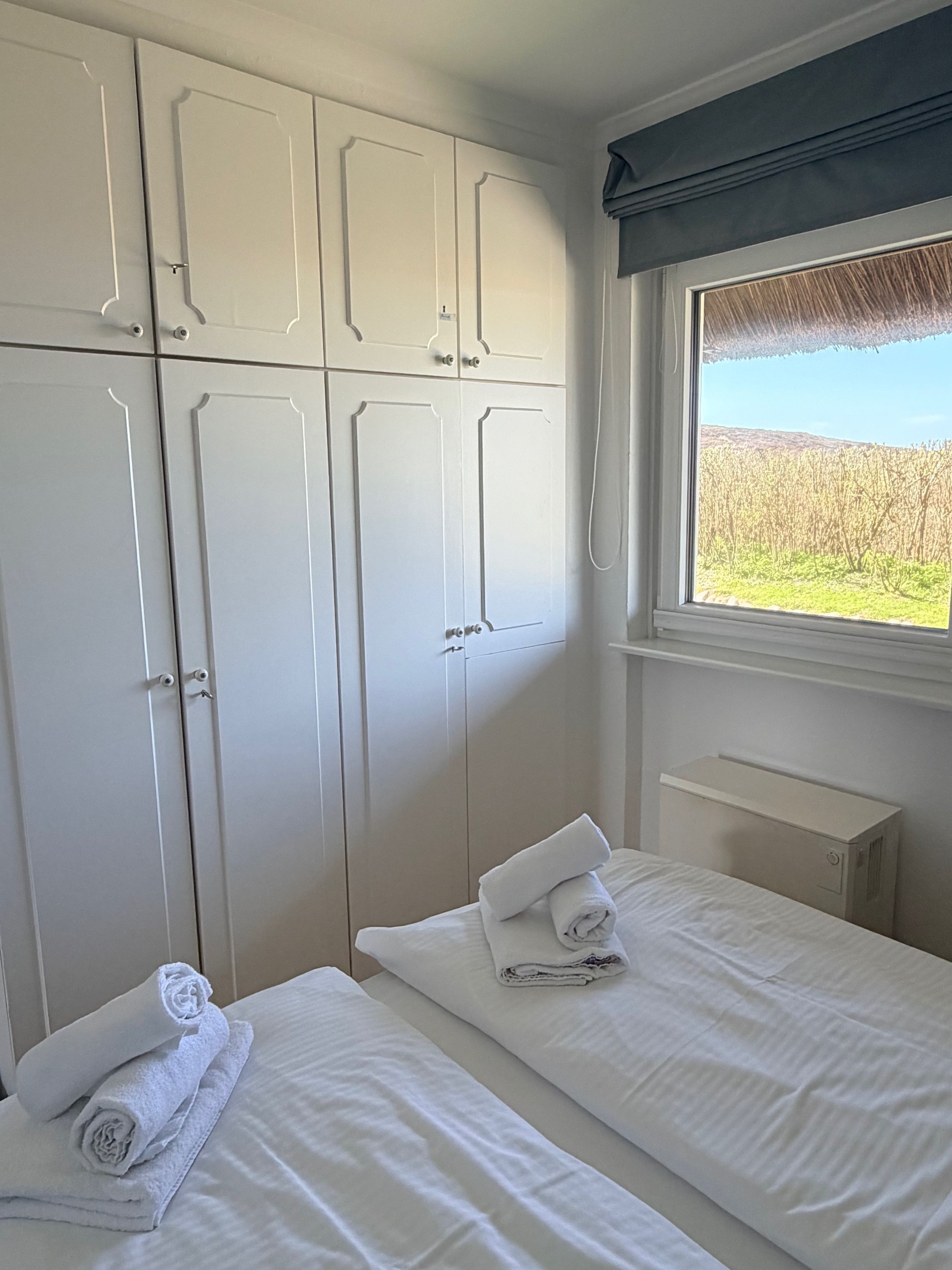 A bedroom with two beds, a wardrobe, a window with a view of a field, and a small bedside table.
