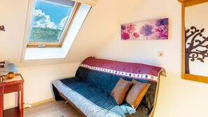 Een gezellige kamer met een schuine balkenlaag, een bed met een beddeken met een patroon, een klein tafeltje met een lamp en een raam met uitzicht op de lucht.