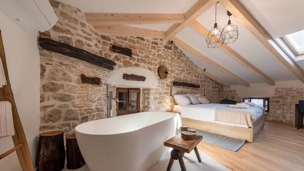 A rustic bedroom with a stone wall, a large bathtub, a wooden bed, and a skylight. - 5 star holiday home in the old town of Bale (Bale)