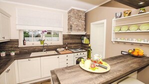 Una cocina con gabinetes blancos, una chimenea de piedra, un ventanal con vistas y una encimera con un frutero y una tabla de cortar.