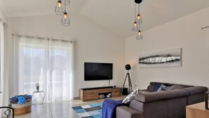Una sala de estar moderna con una televisión de pantalla plana, un sofá, una mesita de centro y un ventanal con cortinas traslúcidas.