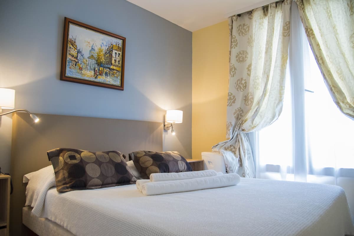 A hotel room with a bed, pillows, a painting on the wall, and a window with curtains.