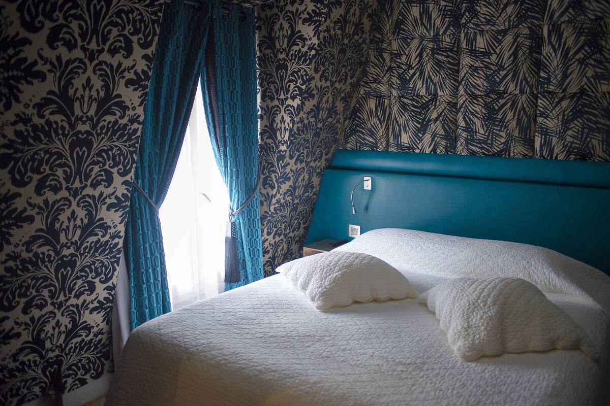 A neatly made bed with white bedding and pillows, a teal headboard, and patterned wallpaper.